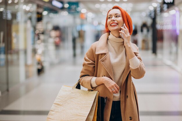 Зрелая женщина с хозяйственными сумками в торговом центре, используя телефон
