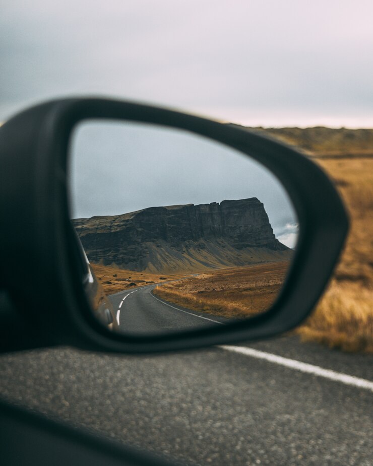 Free Photo meadow surrounded by rocks near the road under a cloudy sky reflecting on a rear-view mirror
