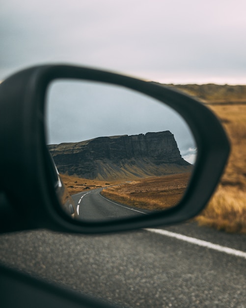 Meadow surrounded by rocks near the road under a cloudy sky reflecting on a Rear-view mirror