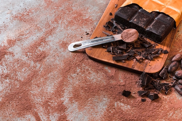 Measuring spoon with cocoa powder and crushed chocolate bar on chopping board