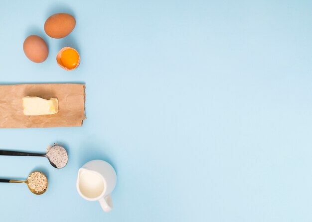 Measuring spoon with oat barn; flour; eggs; butter and milk on blue backdrop