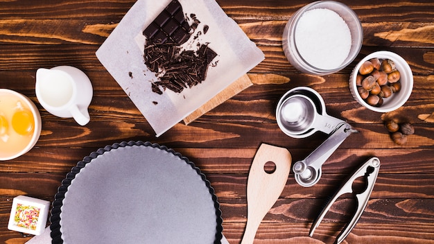 Measuring spoons; chocolate bar; milk; egg yolk; hazelnut and baking dish on wooden textured background