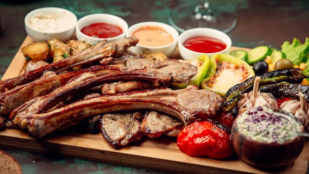 Meat barbecue with grilled vegetables and variety of sauces on a wooden platter.