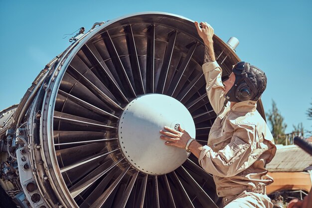 Mechanic in uniform and flying helmet repairing the dismantled airplane turbine in the open air.