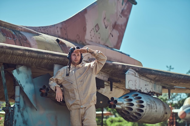 Foto gratuita meccanico in uniforme e casco volante in piedi vicino a un vecchio caccia-intercettore di guerra in un museo a cielo aperto.