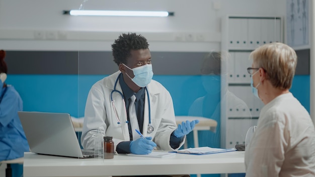 Medic consulting elder patient with face mask and gloves for coronavirus protection at healthcare facility. Young doctor helping senior woman during pandemic at checkup appointment