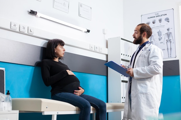 マタニティキャビネットで子供を期待している患者を診察するメディック、妊娠について話している人々。出生前検診で医師の診察やサポートを受けている妊婦。