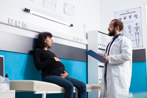 Medic examining patient expecting child in maternity cabinet, people talking about pregnancy. Pregnant woman receiving medical advice and support from doctor at prenatal checkup visit.