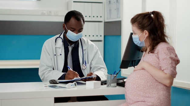 健康診断の訪問でヘルスケアについて話しているフェイスマスクを持つ医者と妊婦。子供を期待している患者との相談、診察予約でメモを取る男性の産科医。