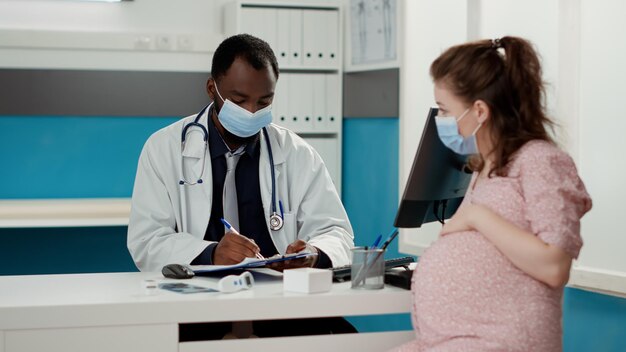 Медик и будущая женщина в маске говорят о медицинском обслуживании во время осмотра. Врач-акушер-мужчина делает записи на консультации с пациентом, ожидающим ребенка, назначение осмотра.