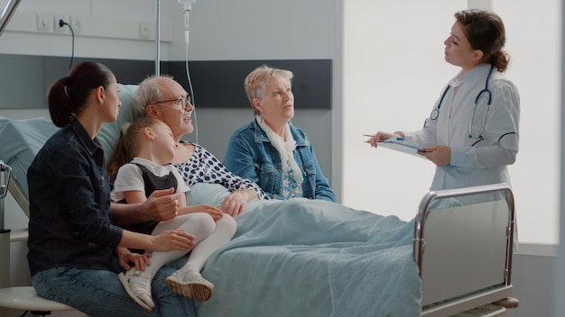 Медик объясняет диагноз пожилому пациенту и его семье в гостях, сидя на больничной койке. Старший мужчина разговаривает с врачом общей практики о лечении и лекарствах, посещает людей.