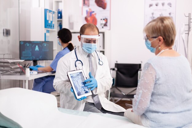 フェイスマスクを身に着けているタブレットPCでX線を指している年配の女性に治療を説明するメディック
