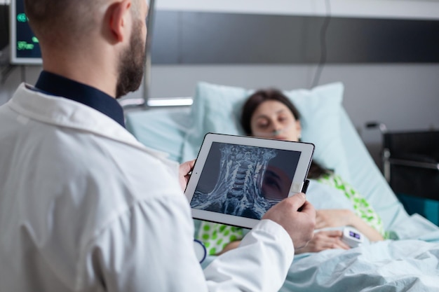 Medic holding digital tablet with throat mri looking at sleeping patient in hospital bed after throat surgery. Woman resting connected to screen measuring vitals after surgical intervention.