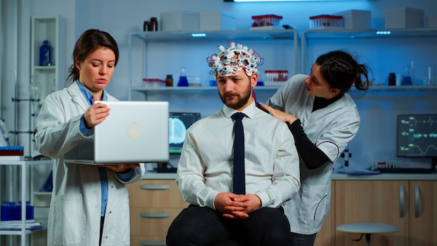Medic in neuroscience working in neurological research laboratory developing brain experiment holding laptop explaning to man brainwave scanning headset side effects of nervous system treamtment