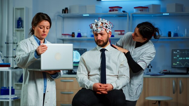 Medic in neuroscience working in neurological research laboratory developing brain experiment holding laptop explaning to man brainwave scanning headset side effects of nervous system treamtment