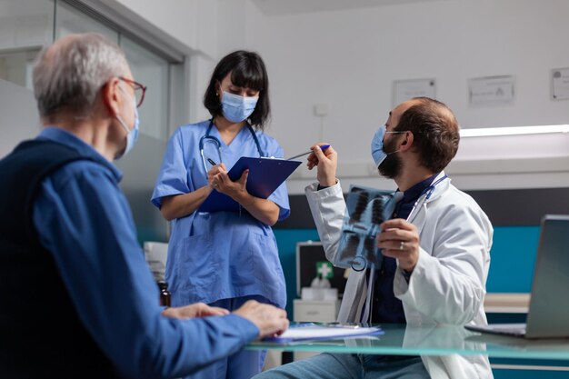 Medic talking to woman nurse and holding x ray results at checkup visit with retired man in office. Doctor explaining radiography diagnosis to patient while he asks assistant to help.