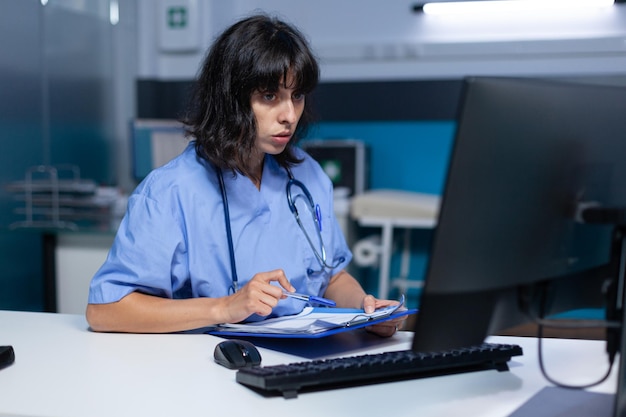 Medical assistant analyzing documents and files on monitor at night. Nurse looking at computer and working for healthcare appointment with checkup papers, doing overtime work at desk.