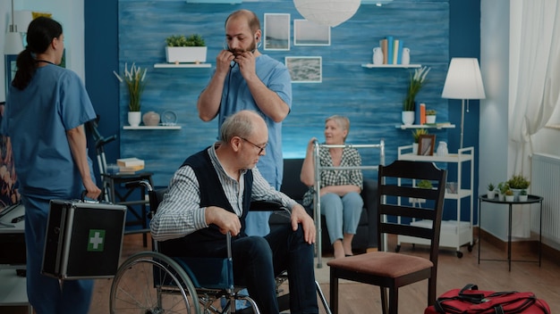 Medical assistant consulting retired man with chronic disability using stethoscope to measure heartbeat and pulse in nursing home. Disabled patient in wheelchair doing checkup with nurse