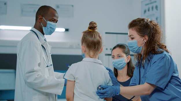 Медицинский помощник консультирует маленького ребенка со стетоскопом в офисе. Женщина-медсестра в униформе проверяет пульс и сердцебиение, ставит диагноз во время пандемии ковида 19.