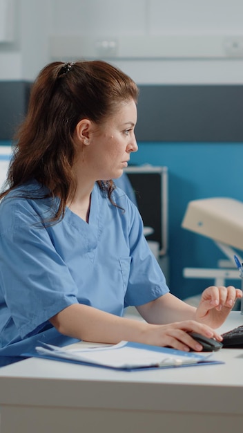 Медицинский ассистент работает на компьютере с информацией о пациенте на столе. Женщина-медсестра использует клавиатуру и монитор в кабинете, проверяя медицинские документы и документы для назначений.