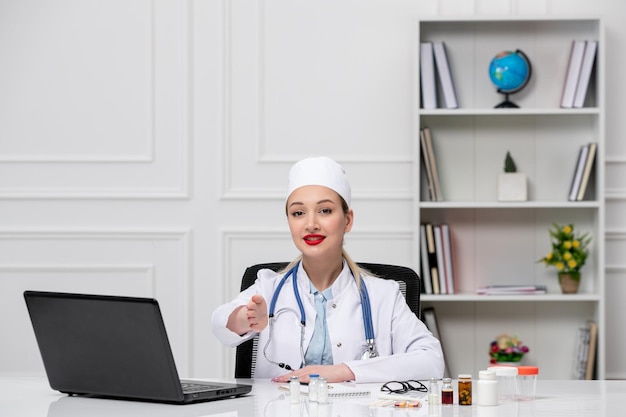 ハンドシェイクを与えるコンピューターと白い白衣と帽子の医療金髪若い医者