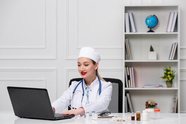 Medical blonde young doctor in white lab coat and hat with computer typing on laptop