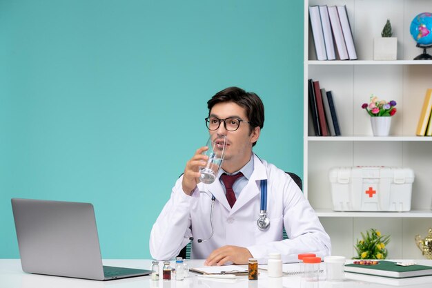 Medical cute handsome doctor in lab coat working remotely on computer drinking water