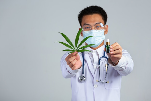 Medical Doctor Holding Cannabis Leaf And Bottle Of Cannabis Oil On White Wall.