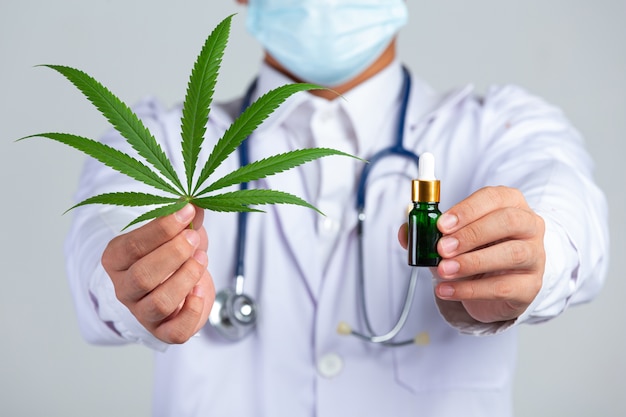 Medical Doctor Holding Cannabis Leaf And Bottle Of Cannabis Oil On White Wall.