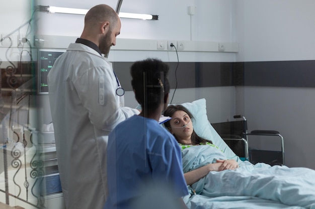 Medical doctor and nurse doing consult in private ward to recovering woman in hospital bed after surgical intervention. Patient with nasal cannula looking at health care professionals.