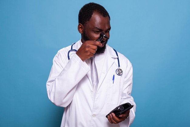 Medical doctor in white lab coat with stethoscope holding otoscope at eye level and otolaryngology kit in hand. Medic in hospital uniform looking through professional device instrument kit.