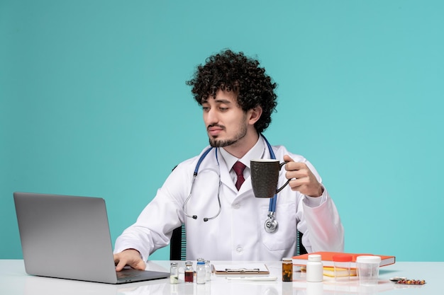 黒のコーヒーカップを保持しているコンピューターに取り組んでいる白衣の若い深刻なハンサムな医師の医療