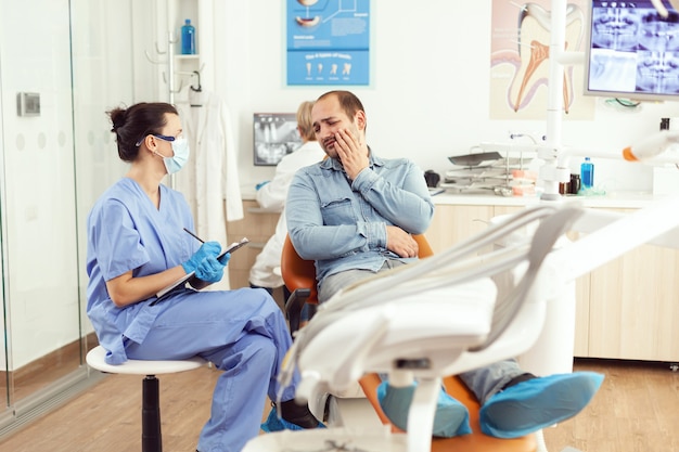 치과 병원 사무실에 앉아 있는 동안 클립보드에 치과 문제를 쓰는 고통스러운 환자를 검사하는 의료 간호사