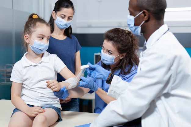 Medical nurse helping african american pediatrician doctor to bandage fractured arm of little kid patient during clinical physiotherapy in hospital office. Team with face mask against covid19