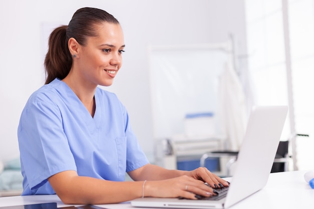 병원 사무실 책상에 앉아 노트북을 사용하여 제복을 입은 의료 간호사. 모니터, 약, 직업, 스크럽을 보고 있는 현대 클리닉에서 컴퓨터를 사용하는 의료 의사.