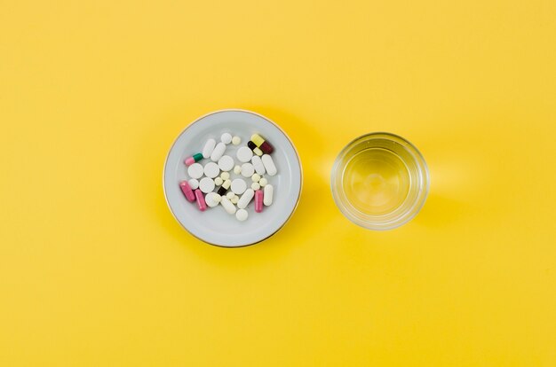 Medical pills in bowl and glass water on yellow background