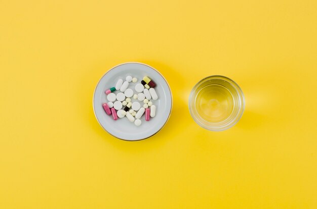 Медицинские таблетки в миску и стакан воды на желтом фоне