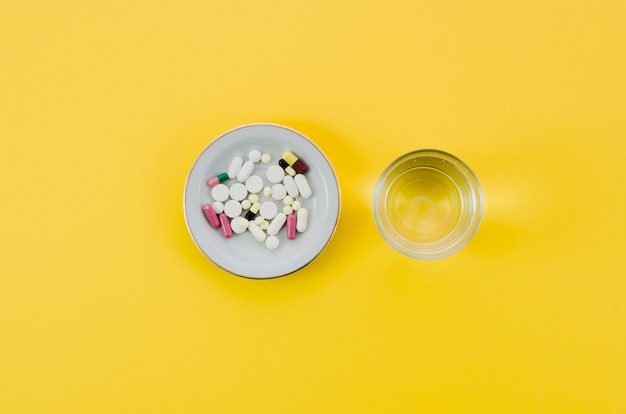 Medical pills in bowl and glass water on yellow background