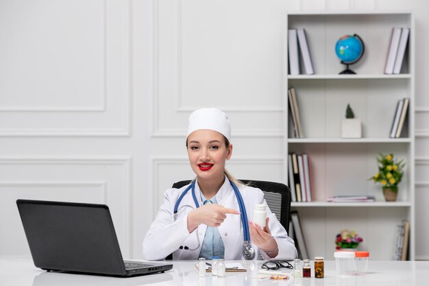 Medical pretty cute doctor in white lab coat and hat with computer pointing at pills