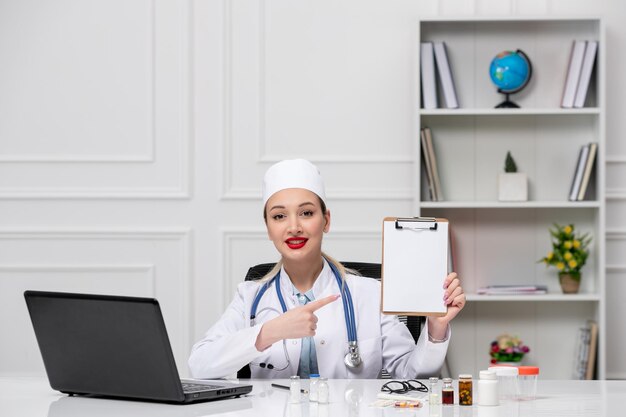 흰색 실험실 코트와 메모를 보여주는 컴퓨터와 모자에 의료 꽤 귀여운 의사
