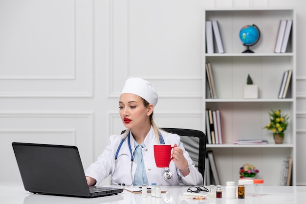 흰색 실험실 코트와 모자와 커피 컵이 있는 컴퓨터를 입은 의료용 예쁜 의사