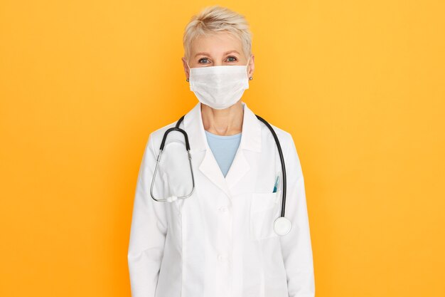 Medical professions, healthcare and prevention concept. Blonde mature senior woman doctor posing isolated wearing white coat and surgical protective mask against infection, virus or bacteria