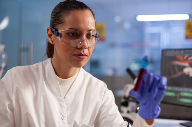 Ученый-медицинский исследователь анализирует образец кувшина крови для испытания развития в химической лаборатории. Профессиональная женщина с лабораторным халатом, очками и перчатками, находящая лечение для здравоохранения