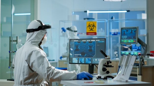장비를 갖춘 실험실에서 PC에 DNA 스캔 이미지 타이핑 작업을 하는 PPE 정장을 입은 의학 과학자. 과학 연구 바이러스 개발을 위한 첨단 화학 도구를 사용하여 백신 진화 조사