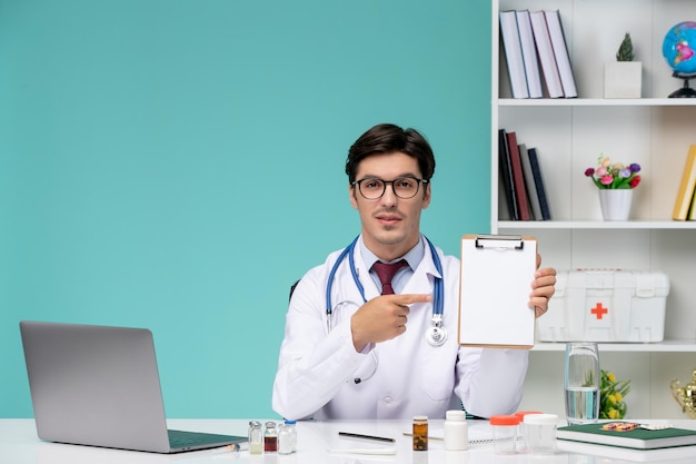 ノートを保持しているコンピューターでリモートで作業している白衣の医療スマート若い医師