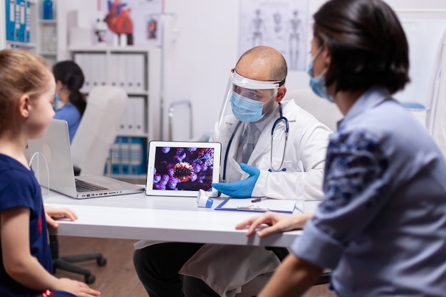 의료 사무실에서 책상에 앉아 태블릿을 사용하여 코로나바이러스를 제시하는 의료 전문가