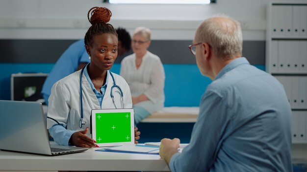 キャビネット内のデジタルタブレットに水平方向の緑色の画面を持ち、高齢患者の孤立した背景を分析する医療専門家。モックアップテンプレートとクロマキー技術を保持している医師。