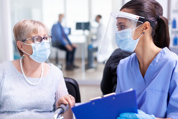 Medical staff with disabled senior woman having a conversation about recovery treatment in hospital waiting area wearing face mask against coronavirus