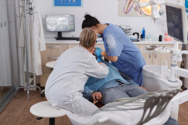 Medical stomatologist team operates patient with caries using dental tools during stomatological examination in dentistry office room. Orthodontist doctor discussing infection treatment with nurse