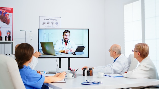 医療チームが会議室で男性専門医とオンライン会議を開催し、クリップボードにメモを取ります。ビデオ通話を使用した患者の治療に関する診断について話し合う医師のグループ